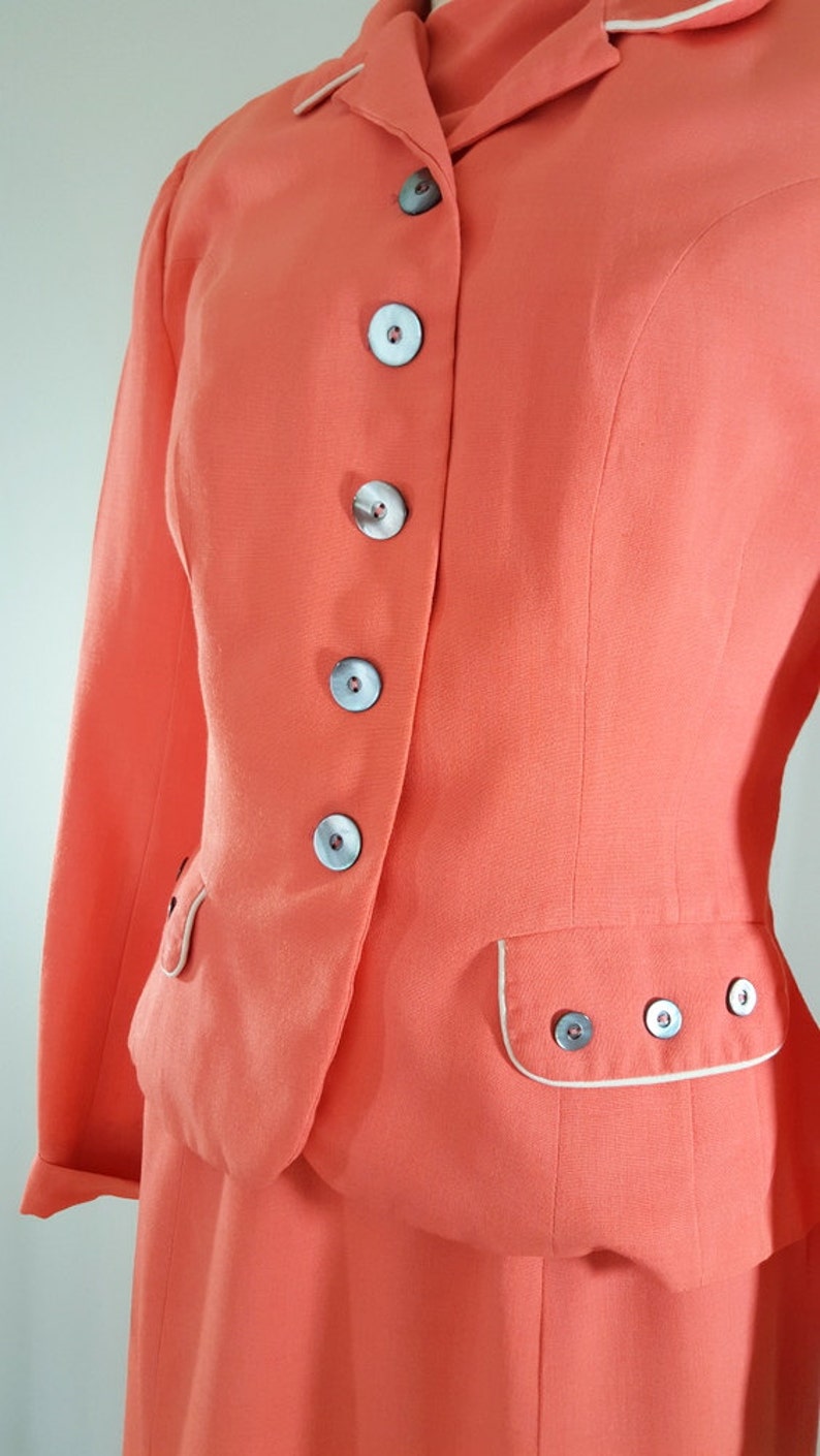 Puede incluir: Un traje vintage de color rosa coral con una chaqueta entallada y una falda. La chaqueta tiene un ribete blanco alrededor del cuello y los bolsillos. La chaqueta tiene cinco botones en la parte delantera, todos con un centro blanco y un anillo exterior gris oscuro.