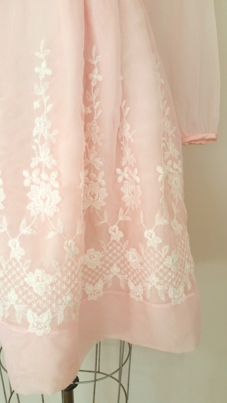 May include: A pink dress with white floral embroidery. The dress has long sleeves and a full skirt.