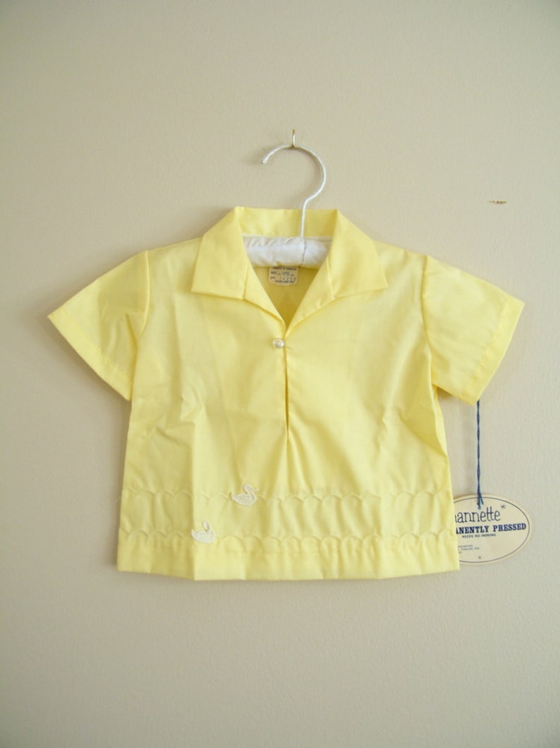 May include: A yellow short-sleeved button-down shirt with a white swan embroidered on the front. The shirt has a scalloped hem and a tag that reads "Annette&reg;  Permanently Pressed".