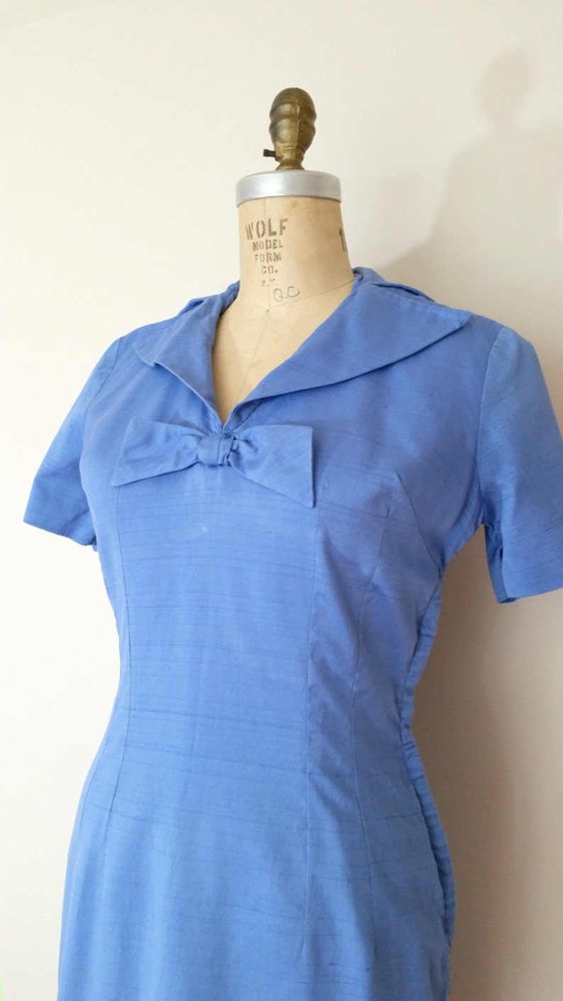 K&ouml;nnte beinhalten: Ein hellblaues Kleid mit Schleife am Ausschnitt und kurzen &Auml;rmeln. Das Kleid befindet sich auf einer Kleiderpuppe.
