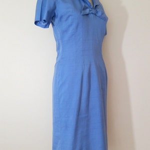 K&ouml;nnte beinhalten: Ein hellblaues Kleid mit einer Schleife am Ausschnitt und kurzen &Auml;rmeln. Das Kleid ist figurbetont und hat einen geraden Rock.