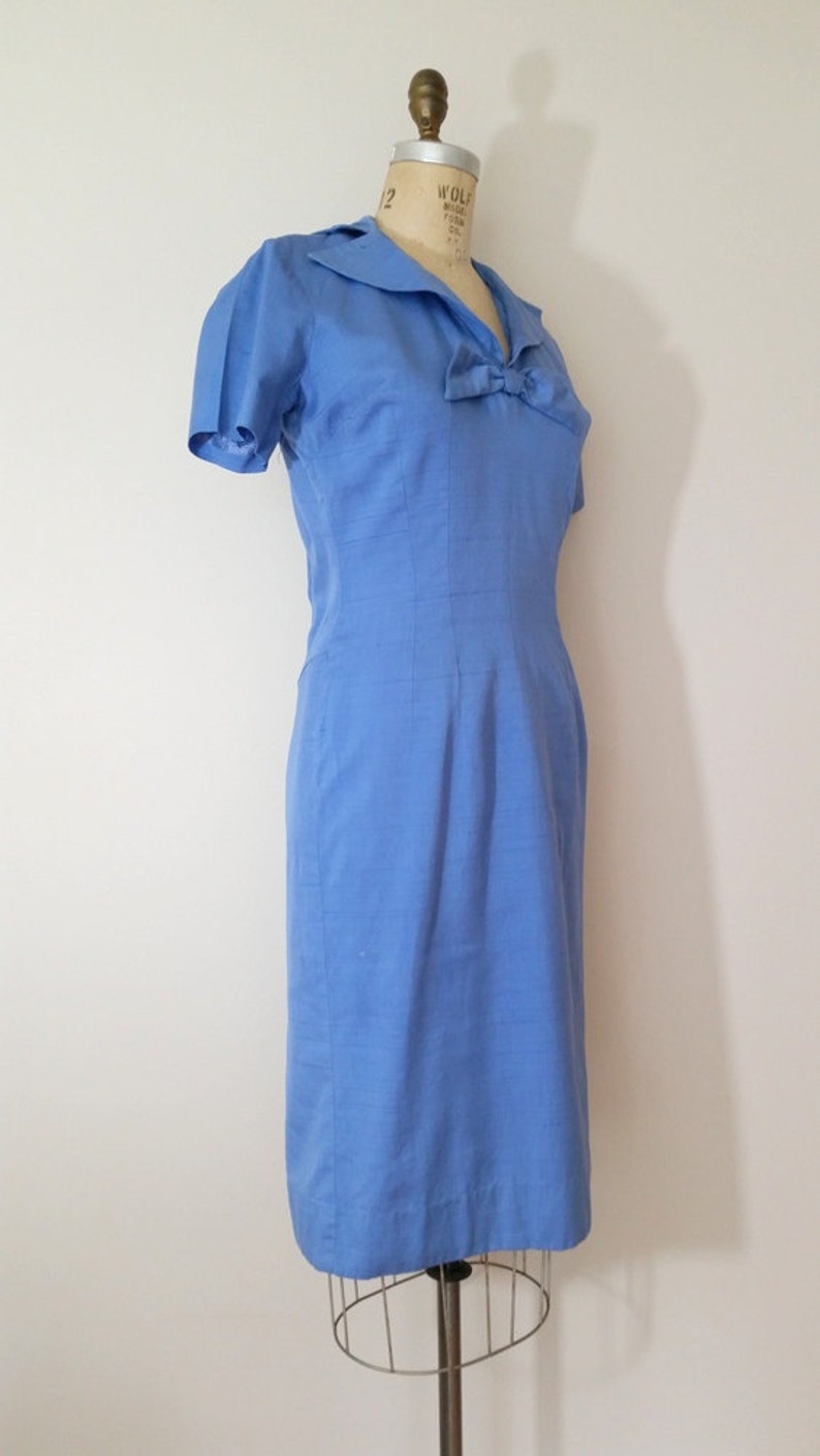 K&ouml;nnte beinhalten: Ein hellblaues Kleid mit einer Schleife am Ausschnitt und kurzen &Auml;rmeln. Das Kleid ist figurbetont und hat einen geraden Rock.