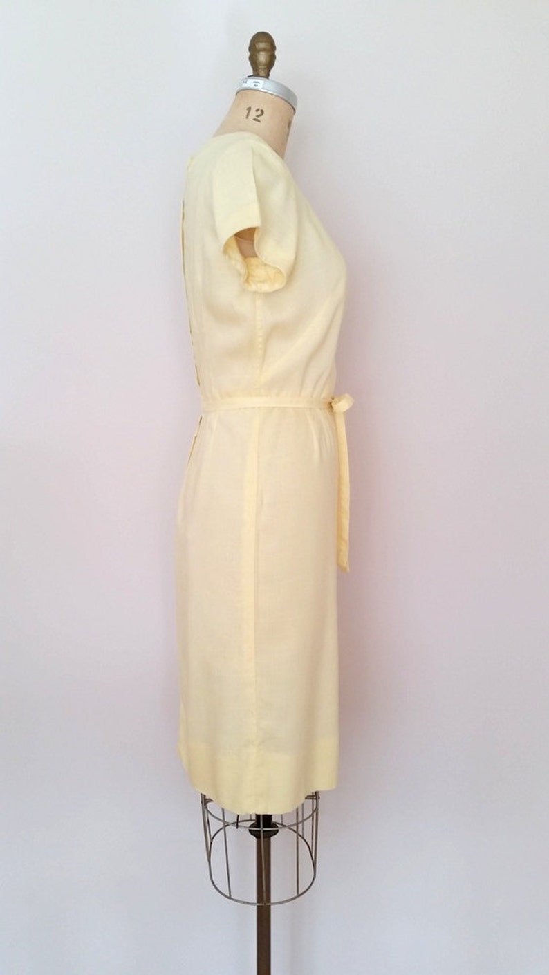 May include: A pale yellow sleeveless dress with a sash tie at the waist. The dress is on a dress form.