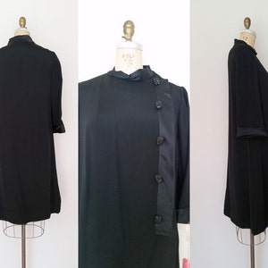 K&ouml;nnte beinhalten: Ein schwarzes Kleid mit Stehkragen und langem, flie&szlig;endem Rock. Das Kleid hat einen Satinbesatz und Kn&ouml;pfe an der Seite.