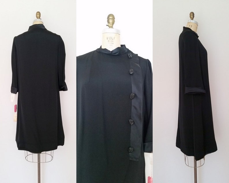 K&ouml;nnte beinhalten: Ein schwarzes Kleid mit Stehkragen und langem, flie&szlig;endem Rock. Das Kleid hat einen Satinbesatz und Kn&ouml;pfe an der Seite.