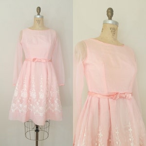 May include: A pink vintage dress with long sheer sleeves and a white floral embroidered design. The dress has a fitted bodice and a full skirt with a bow at the waist.