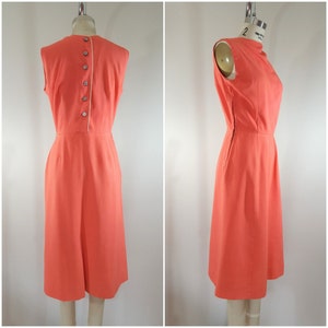 Puede incluir: Un vestido coral sin mangas con cierre de botones en la espalda. El vestido tiene una silueta simple y ajustada y un largo hasta la rodilla.