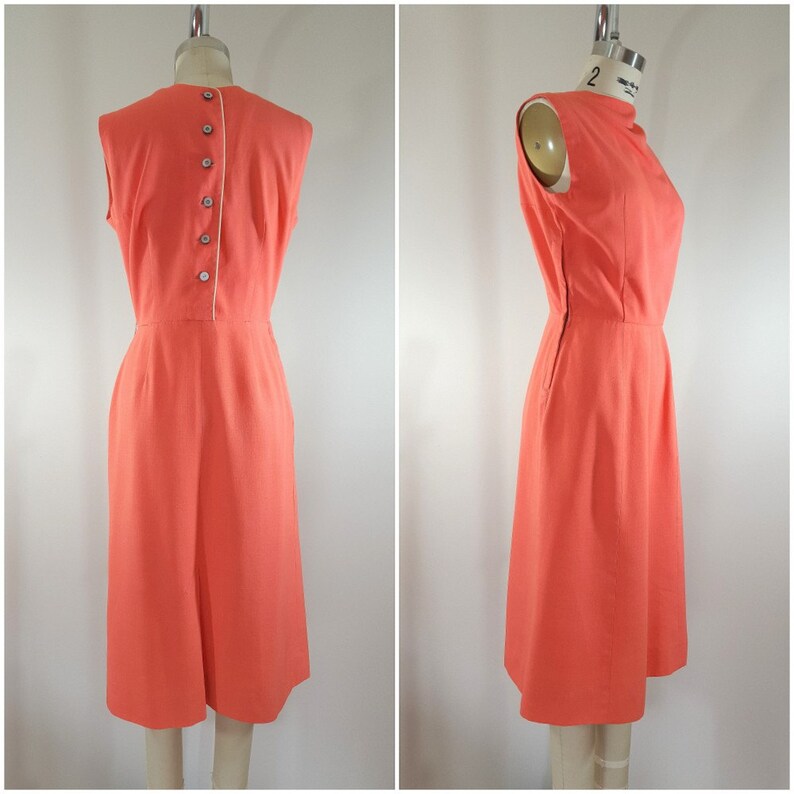 Puede incluir: Un vestido coral sin mangas con cierre de botones en la espalda. El vestido tiene una silueta simple y ajustada y un largo hasta la rodilla.