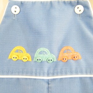 Op de afbeelding: Blauwe corduroy overall met witte bies en drie geborduurde auto's in geel, groen en oranje.