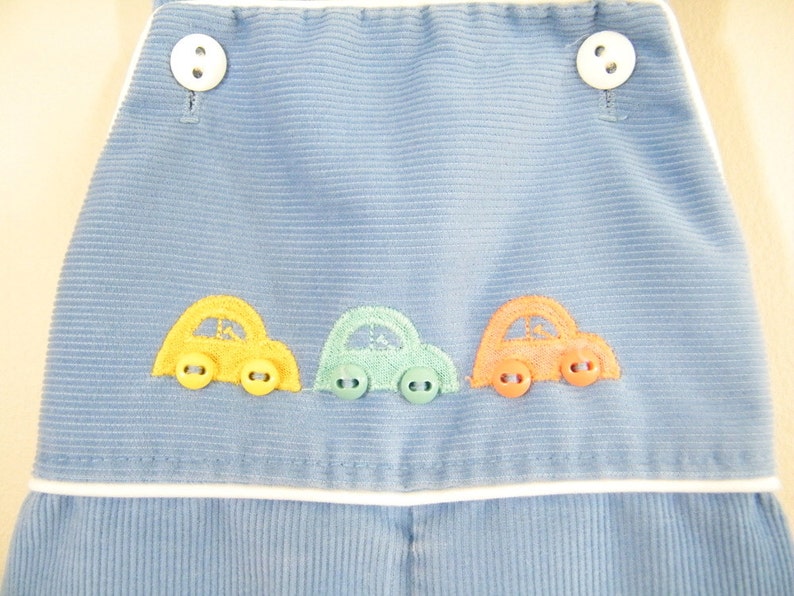 Op de afbeelding: Blauwe corduroy overall met witte bies en drie geborduurde auto's in geel, groen en oranje.