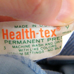 Op de afbeelding: Een wit label met groene en oranje tekst die "Health-tex&reg; Permanent Press Machine Wash and Dry with Like Colors Medium Settings - No Bleach 9 mos" zegt.