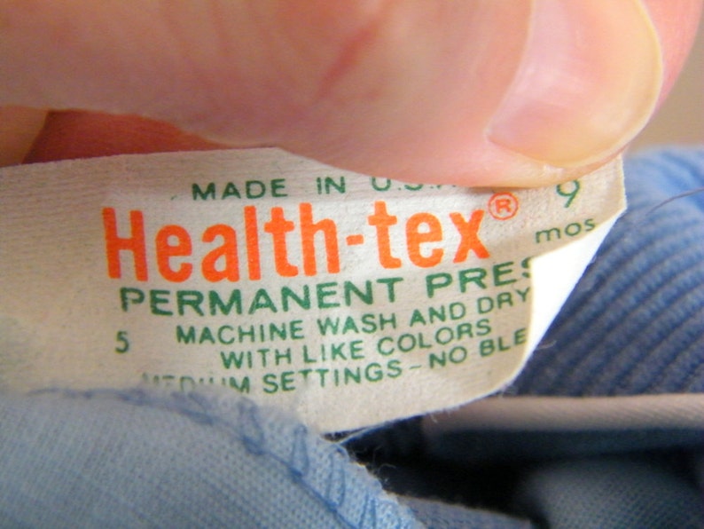 Op de afbeelding: Een wit label met groene en oranje tekst die "Health-tex&reg; Permanent Press Machine Wash and Dry with Like Colors Medium Settings - No Bleach 9 mos" zegt.