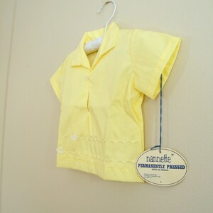 May include: A yellow short-sleeved shirt with a white embroidered design. The shirt is hanging on a hanger and has a tag that reads "nannette* PERMANENTLY PRESSED NEEDS NO IRONING".