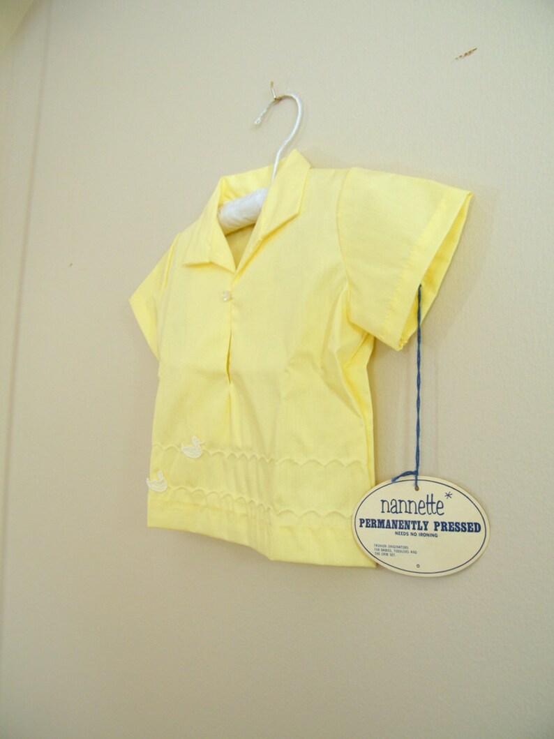 May include: A yellow short-sleeved shirt with a white embroidered design. The shirt is hanging on a hanger and has a tag that reads "nannette* PERMANENTLY PRESSED NEEDS NO IRONING".