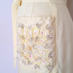May include: A pale yellow linen dress with a large embroidered floral pocket detail. The embroidery features yellow, white, and peach flowers with green leaves.