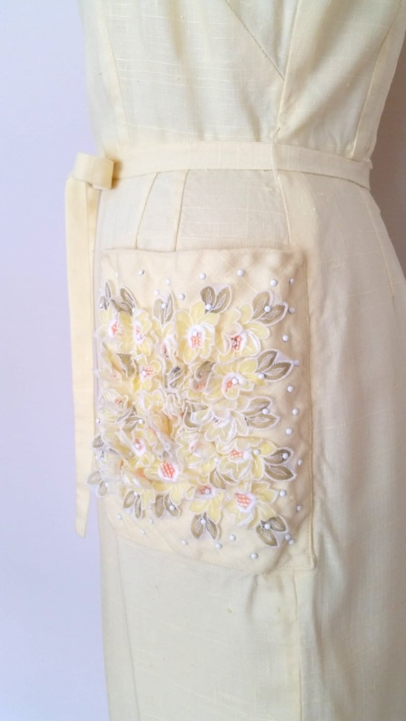 May include: A pale yellow linen dress with a large embroidered floral pocket detail. The embroidery features yellow, white, and peach flowers with green leaves.