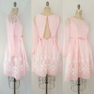 May include: A pink dress with long sheer sleeves and a white floral embroidered design. The dress has a low back with a zipper closure.