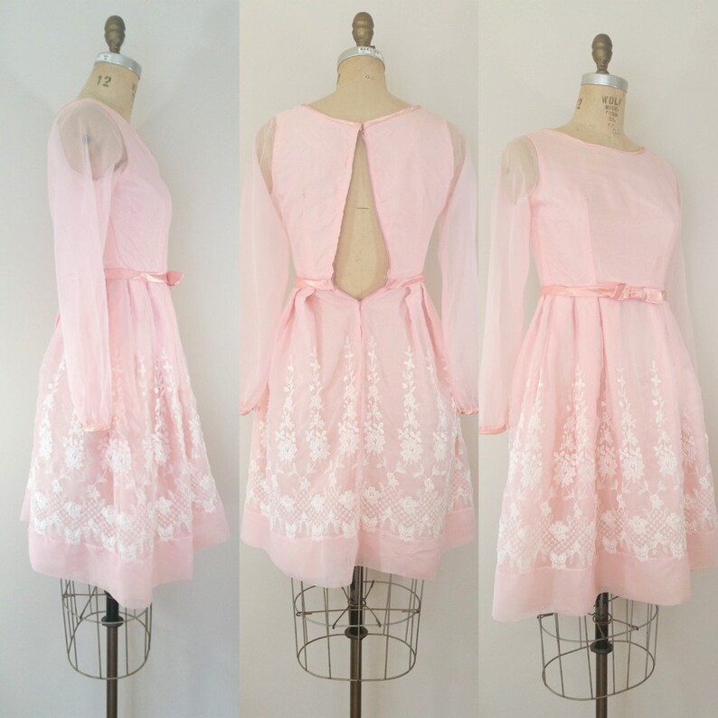 May include: A pink dress with long sheer sleeves and a white floral embroidered design. The dress has a low back with a zipper closure.