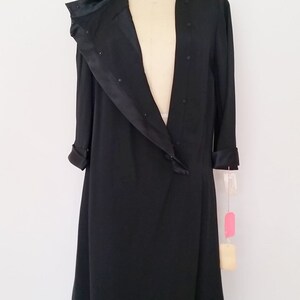 K&ouml;nnte beinhalten: Ein schwarzes Satin-Kleid mit langem, flie&szlig;endem Rock und einem Schalkragen. Das Kleid befindet sich auf einer Schaufensterpuppe und hat ein Etikett an der &Auml;rmel.