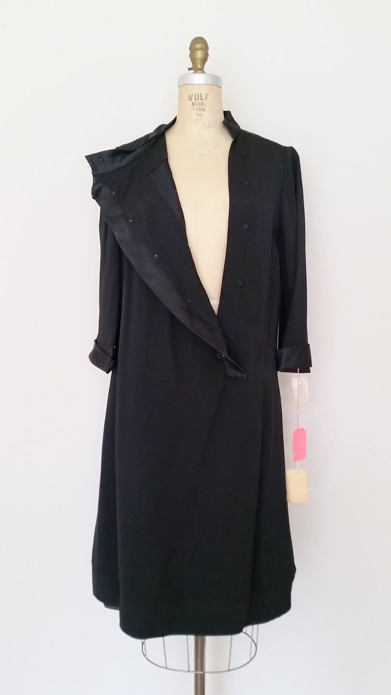 K&ouml;nnte beinhalten: Ein schwarzes Satin-Kleid mit langem, flie&szlig;endem Rock und einem Schalkragen. Das Kleid befindet sich auf einer Schaufensterpuppe und hat ein Etikett an der &Auml;rmel.
