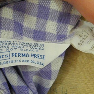 May include: A white and purple gingham fabric label with the text "NO IRONING IF TUMBLE DRIED AND REMOVED IMMEDIATELY MACHINE WASH - WARM DO NOT BLEACH Sears PERMA-PREST SEARS, ROEBUCK AND CO., U.S.A." and a white tag with the text "E-2 8252/7/4 SIZE 12 100 %POLYES KNIT".