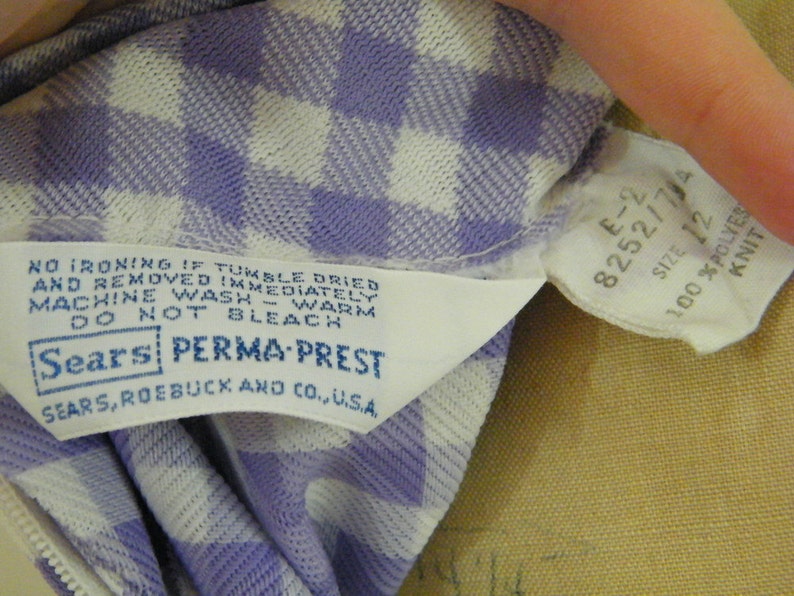 May include: A white and purple gingham fabric label with the text "NO IRONING IF TUMBLE DRIED AND REMOVED IMMEDIATELY MACHINE WASH - WARM DO NOT BLEACH Sears PERMA-PREST SEARS, ROEBUCK AND CO., U.S.A." and a white tag with the text "E-2 8252/7/4 SIZE 12 100 %POLYES KNIT".