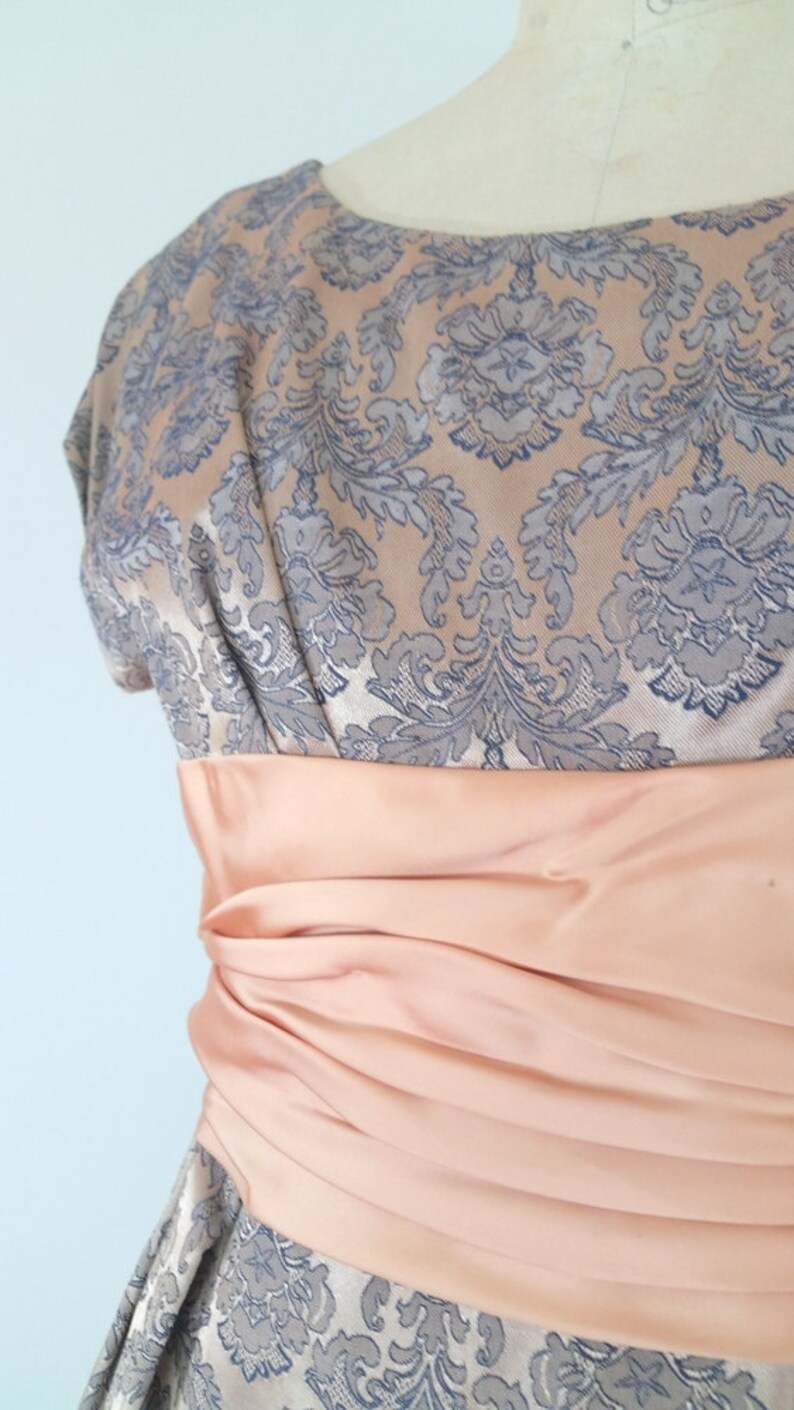 May include: A vintage dress with a floral pattern in shades of gray and peach. The dress has a fitted bodice and a full skirt. A peach-colored satin sash is tied around the waist.