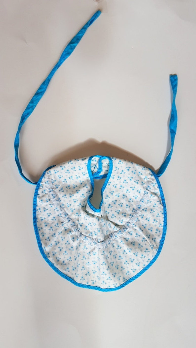 May include: A white baby bib with a blue floral pattern and blue trim. The bib has two blue ties.