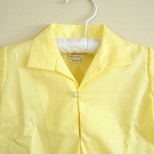May include: A yellow short-sleeved shirt with a button-down collar and a single button closure. The shirt has a label that reads "SIZE: sm-m (10-12)  STY: 14239 MADE IN U.S.A."