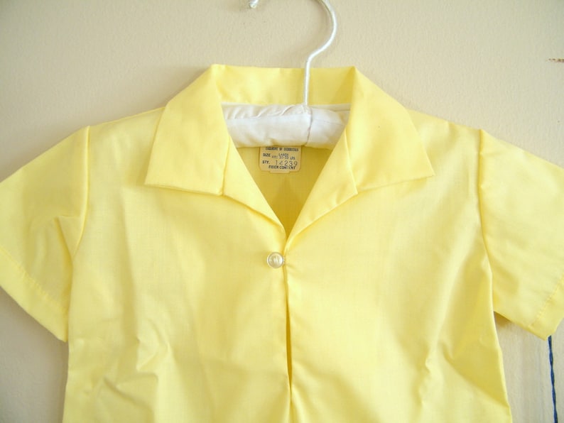 May include: A yellow short-sleeved shirt with a button-down collar and a single button closure. The shirt has a label that reads "SIZE: sm-m (10-12)  STY: 14239 MADE IN U.S.A."