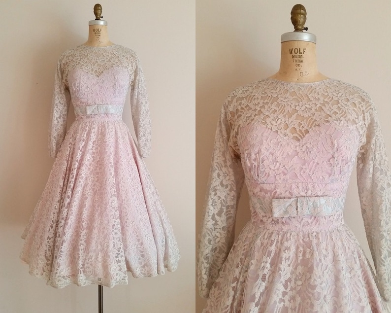 May include: A vintage pink lace dress with a fitted bodice and a full skirt. The dress has long sleeves and a white satin ribbon at the waist.