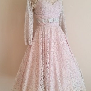 May include: A pale pink lace dress with long sleeves and a fitted bodice. The dress has a full skirt and a silver ribbon tied at the waist.