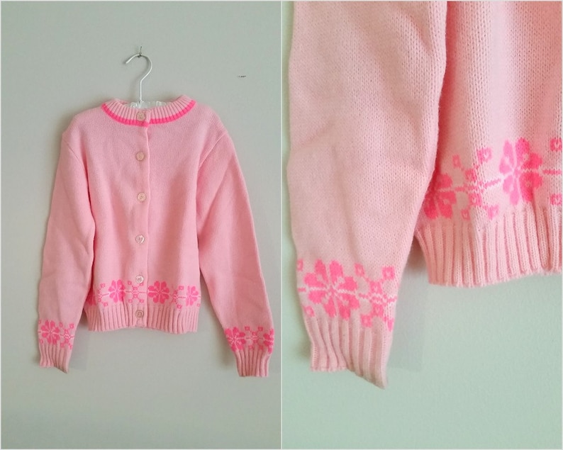 Puede incluir: Un c&aacute;rdigan de punto rosa con cierre de botones y bordado floral rosa. El c&aacute;rdigan tiene mangas largas y un dobladillo de canal&eacute;.