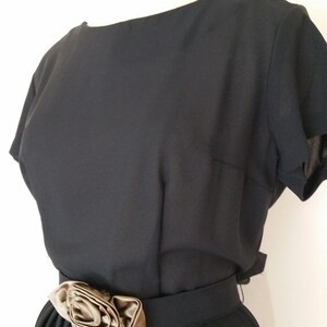 May include: A black dress with a short sleeve and a brown satin flower accent on the waist.