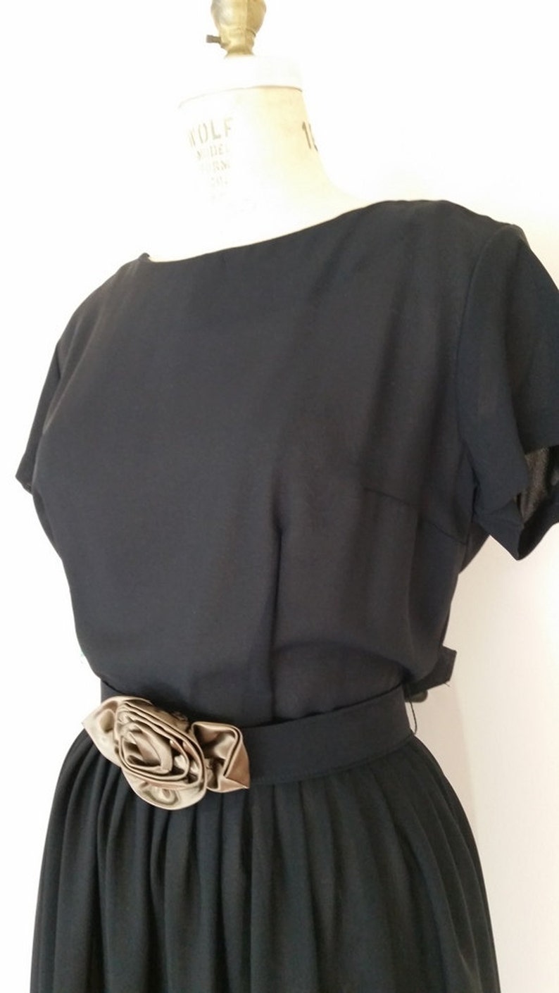 May include: A black dress with a short sleeve and a brown satin flower accent on the waist.