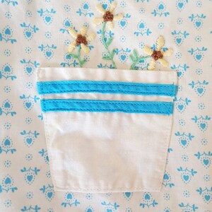 May include: A white fabric pocket with two blue stripes on the top edge. The pocket is embroidered with a small bouquet of yellow flowers with brown centres.