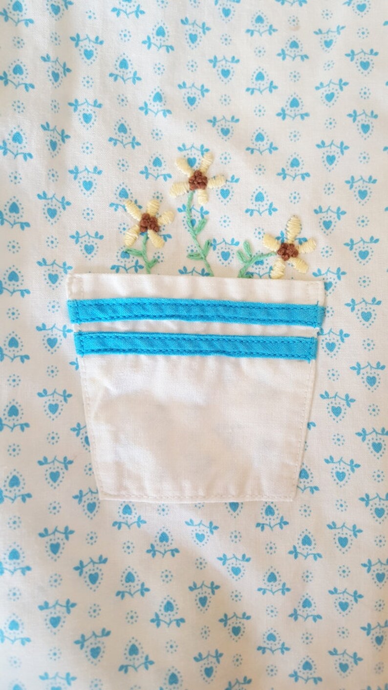 May include: A white fabric pocket with two blue stripes on the top edge. The pocket is embroidered with a small bouquet of yellow flowers with brown centres.