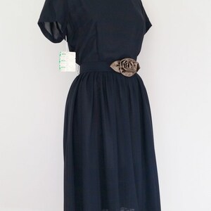 May include: A black vintage dress with a bateau neckline and short sleeves. The dress has a fitted bodice and a full skirt. A brown satin rose embellishment is attached to the black belt.