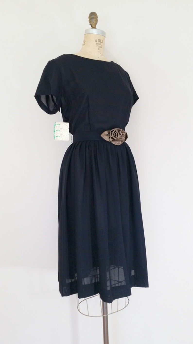 May include: A black vintage dress with a bateau neckline and short sleeves. The dress has a fitted bodice and a full skirt. A brown satin rose embellishment is attached to the black belt.