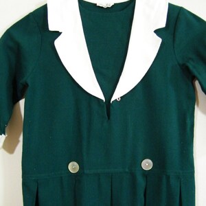 Puede incluir: Un vestido verde oscuro con cuello blanco y dos botones. El vestido tiene un escote en V y una falda plisada.