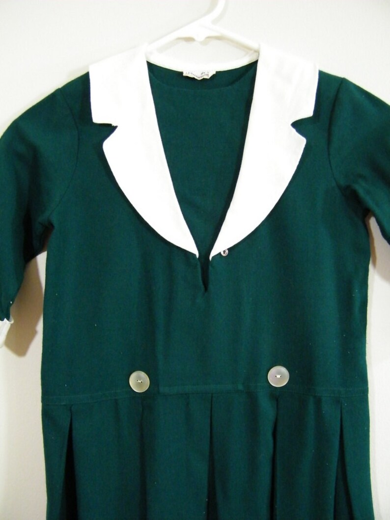 Puede incluir: Un vestido verde oscuro con cuello blanco y dos botones. El vestido tiene un escote en V y una falda plisada.