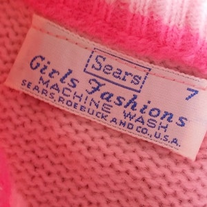Puede incluir: Un su&eacute;ter de punto rosa con una etiqueta blanca que dice "Girls Fashions [Sears] Machine Wash Sears, Roebuck and Co., U.S.A. 7"