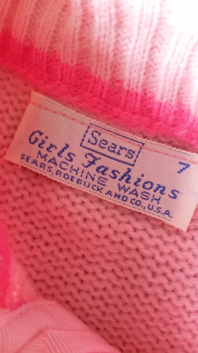 Puede incluir: Un su&eacute;ter de punto rosa con una etiqueta blanca que dice "Girls Fashions [Sears] Machine Wash Sears, Roebuck and Co., U.S.A. 7"