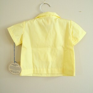May include: A yellow short-sleeved button-down shirt hanging on a white hanger. The shirt has a collar and a tag attached to the bottom.