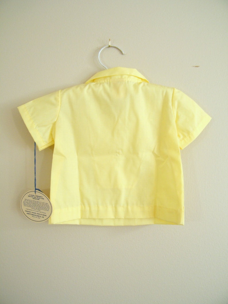 May include: A yellow short-sleeved button-down shirt hanging on a white hanger. The shirt has a collar and a tag attached to the bottom.