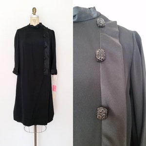 K&ouml;nnte beinhalten: Ein schwarzes Kleid mit hohem Kragen und langen &Auml;rmeln. Das Kleid hat eine diagonale Naht vorne mit Kn&ouml;pfen auf einer Seite. Die Kn&ouml;pfe sind mit Strasssteinen verziert.