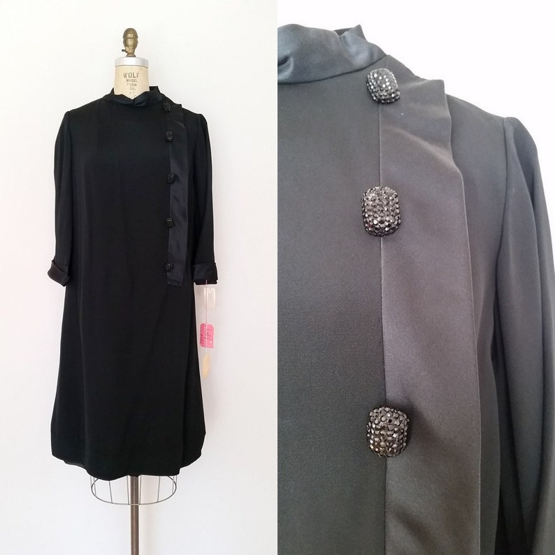 K&ouml;nnte beinhalten: Ein schwarzes Kleid mit hohem Kragen und langen &Auml;rmeln. Das Kleid hat eine diagonale Naht vorne mit Kn&ouml;pfen auf einer Seite. Die Kn&ouml;pfe sind mit Strasssteinen verziert.