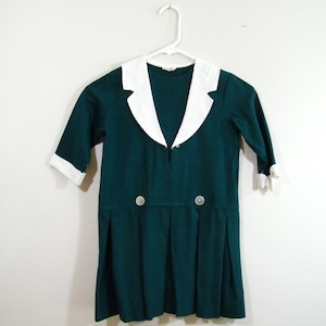 Puede incluir: Un vestido verde oscuro con un cuello blanco Peter Pan y dos botones en la parte delantera. El vestido tiene una falda plisada y mangas cortas.