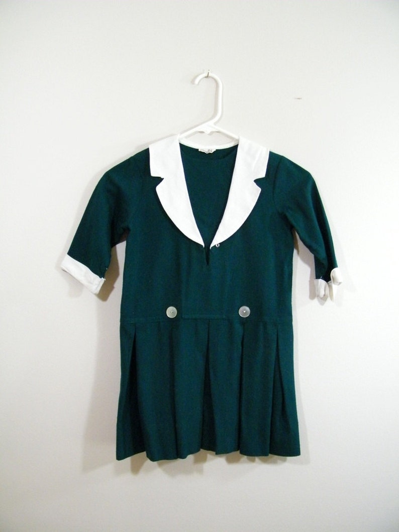 Puede incluir: Un vestido verde oscuro con un cuello blanco Peter Pan y dos botones en la parte delantera. El vestido tiene una falda plisada y mangas cortas.