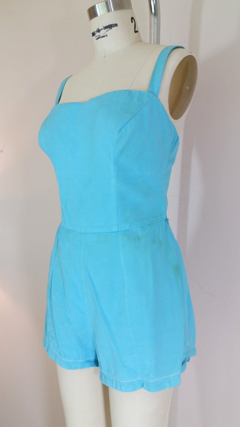 May include: A light blue sleeveless jumpsuit with a square neckline and a fitted bodice. The jumpsuit has a wide leg and a hem that falls just above the knee.